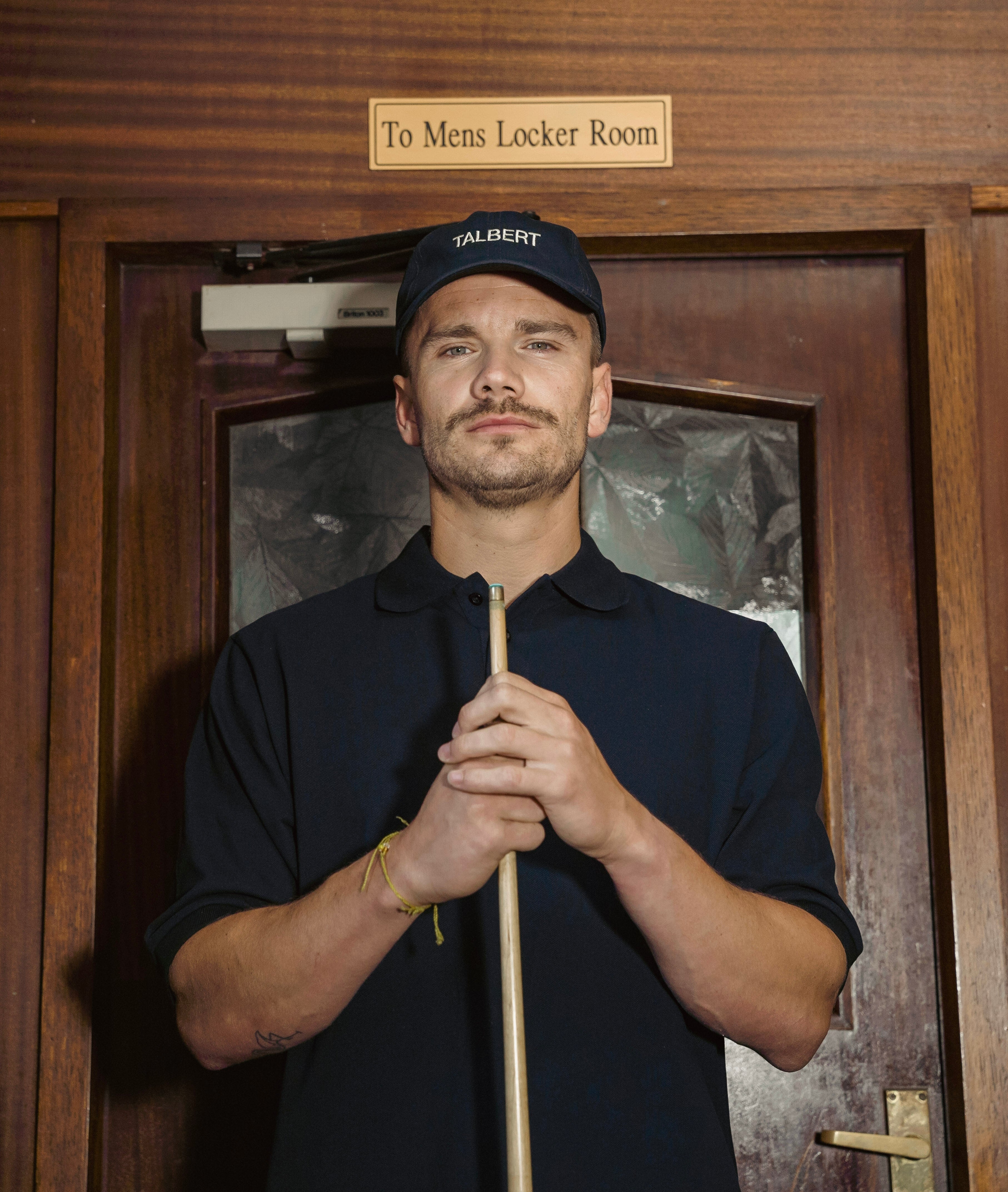 Man holding a pool cue in front of a wooden door labeled 'To Mens Locker Room'.